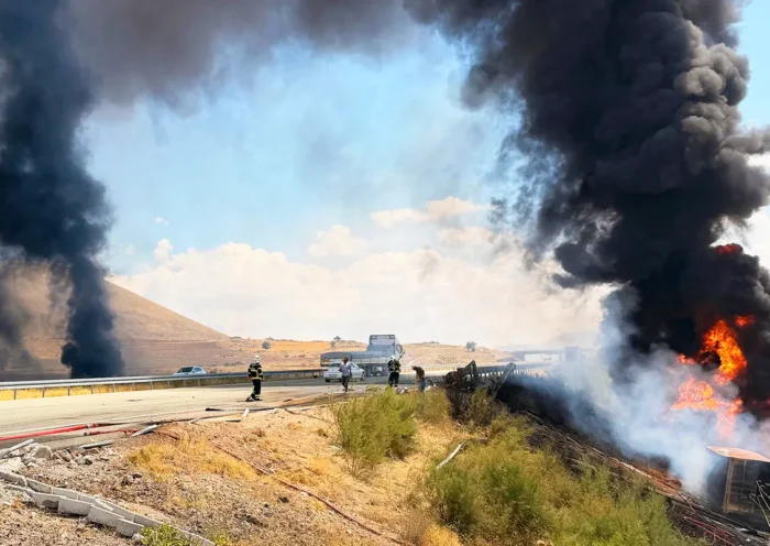varios bomberos junto a una autopista y un camión ardiendo en medio de grandes columnas de humo