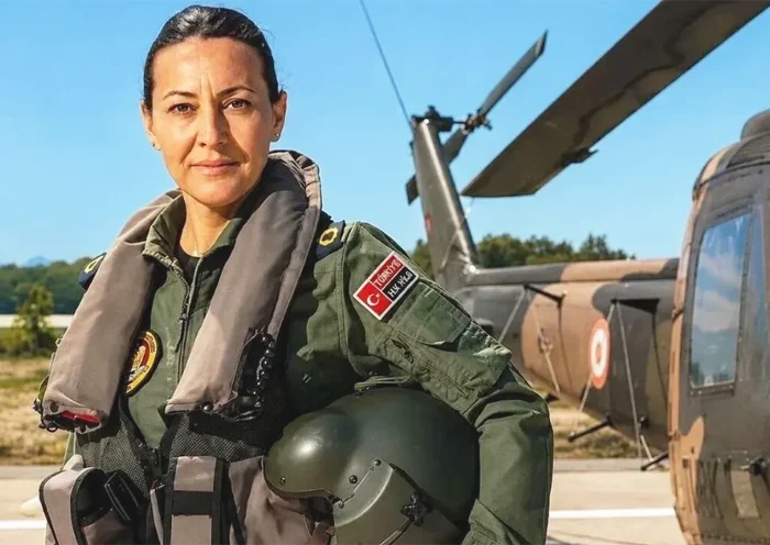 una mujer mirando a la cámara vistiendo un traje de piloto de las Fuerzas Aéreas Turcas posa con un casco bajo el brazo y junto a un helicóptero
