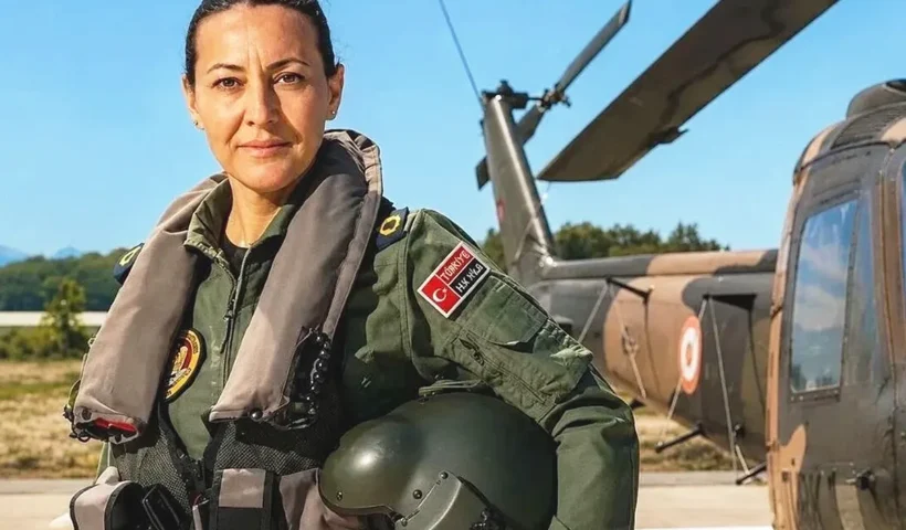 una mujer mirando a la cámara vistiendo un traje de piloto de las Fuerzas Aéreas Turcas posa con un casco bajo el brazo y junto a un helicóptero
