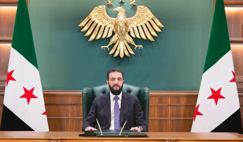 el presidente sirio Ahmad al-Sharaa hablando desde su despacho con la bandera revolucionaria siria y el escudo del país de fondo