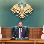 Syrian President Ahmad al-Sharaa speaking from his office with the Syrian revolutionary flag and the country’s coat of arms in the background