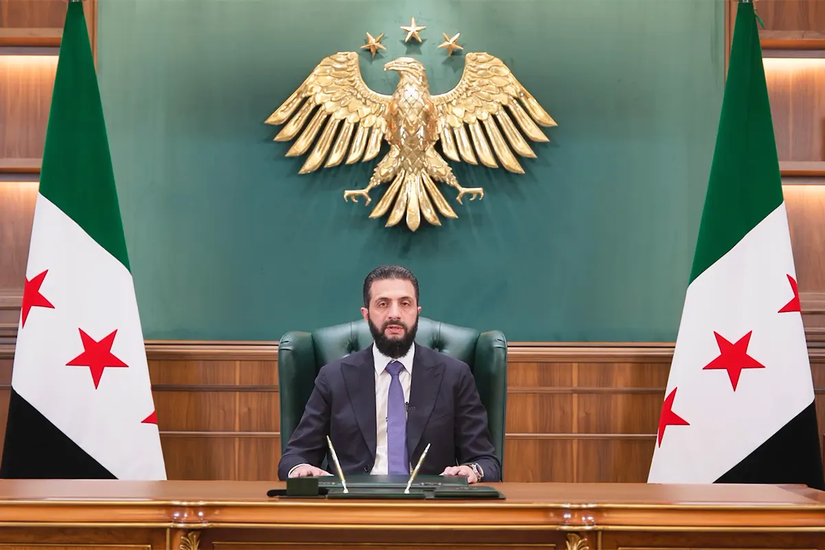 Syrian President Ahmad al-Sharaa speaking from his office with the Syrian revolutionary flag and the country’s coat of arms in the background