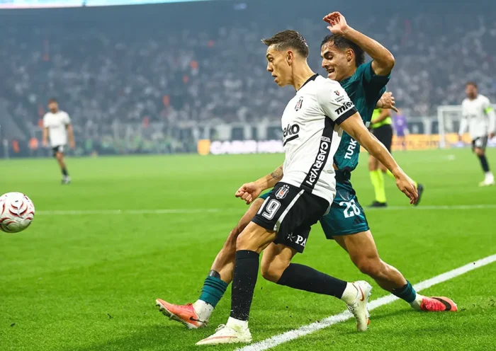 A Beşiktaş player and a Kocaelispor player battling for the ball by the touchline in a packed stadium at night