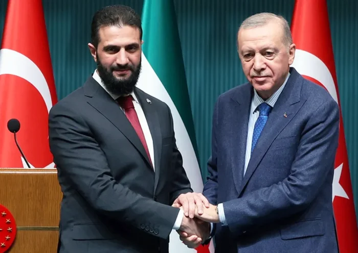 una imagen de Al-Sharaa estrechando sus manos con Erdoğan con las banderas de Turquía y Siria de fondo
