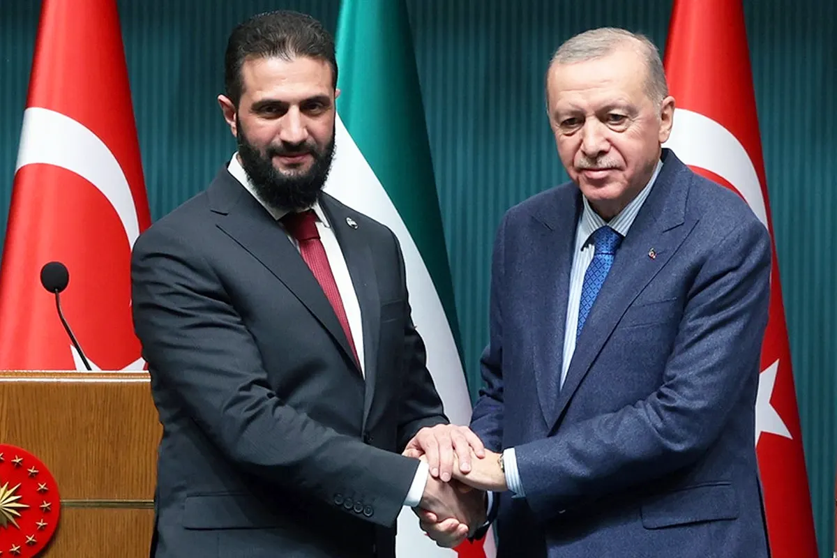 una imagen de Al-Sharaa estrechando sus manos con Erdoğan con las banderas de Turquía y Siria de fondo