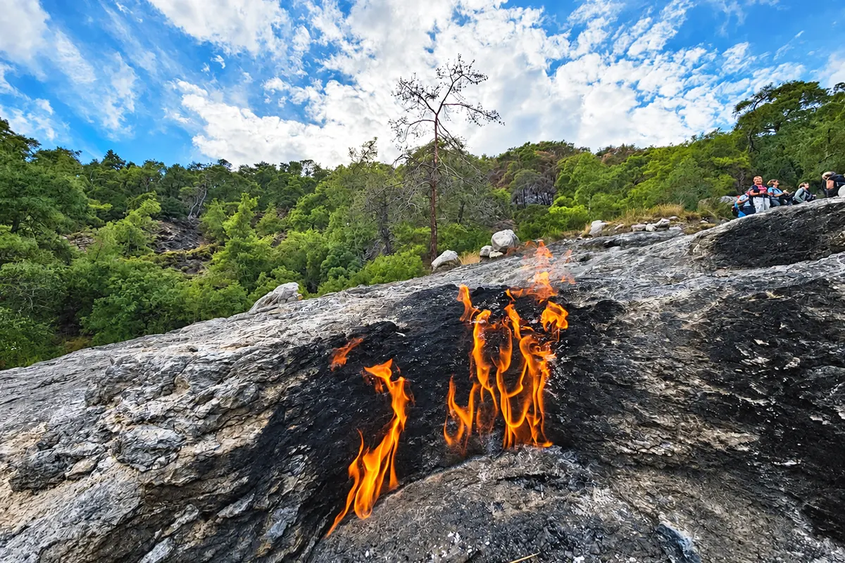 Flames emerging from between the rocks at Yanartaş, with a group of walkers watching the phenomenon and a forest in the background